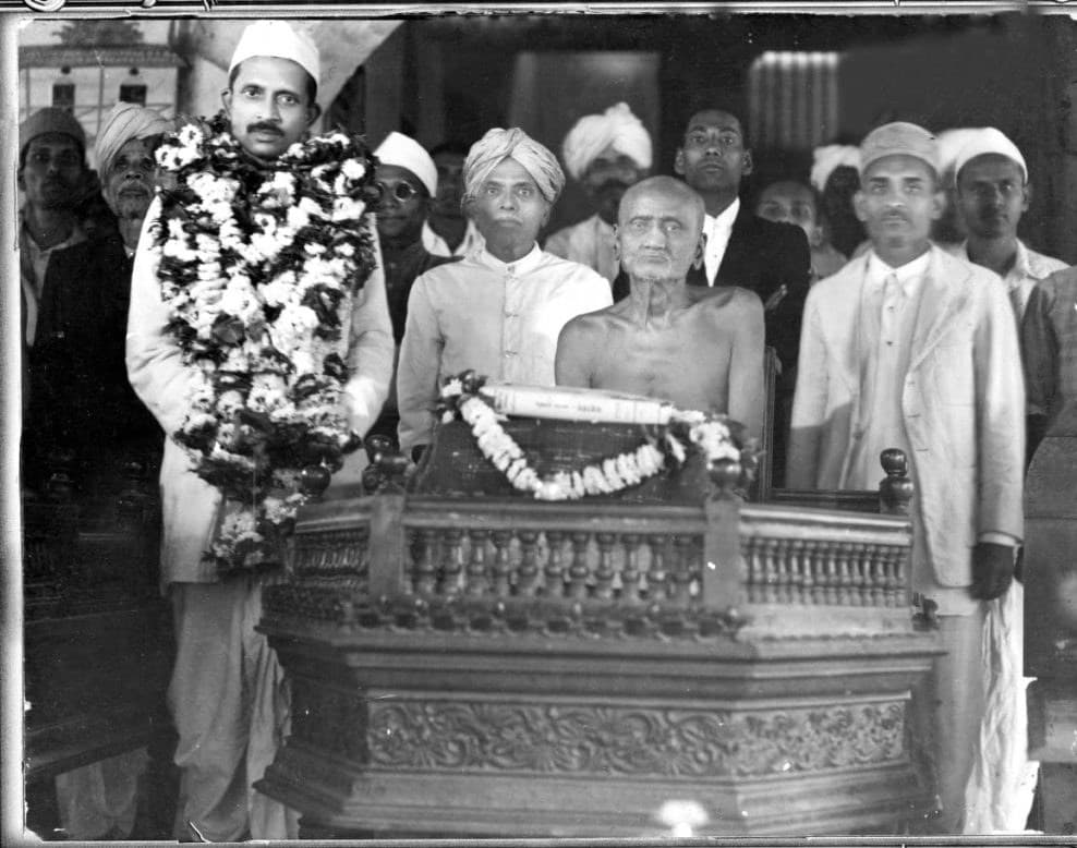 Black-and-white group photograph: the Acharya seated behind a carved stand that bears a garlanded scripture, with a garlanded man and others standing around him.