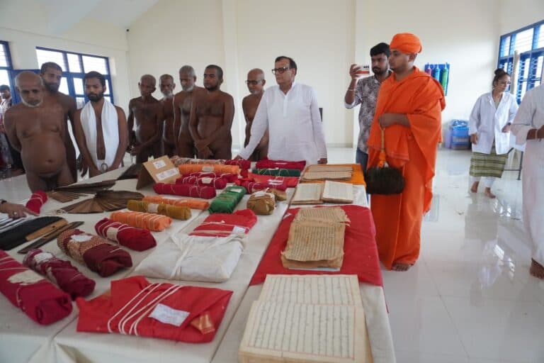 Palm-leaf and paper manuscripts laid out on red cloth at Shravanabelagola, with monks and Foundation members looking on.