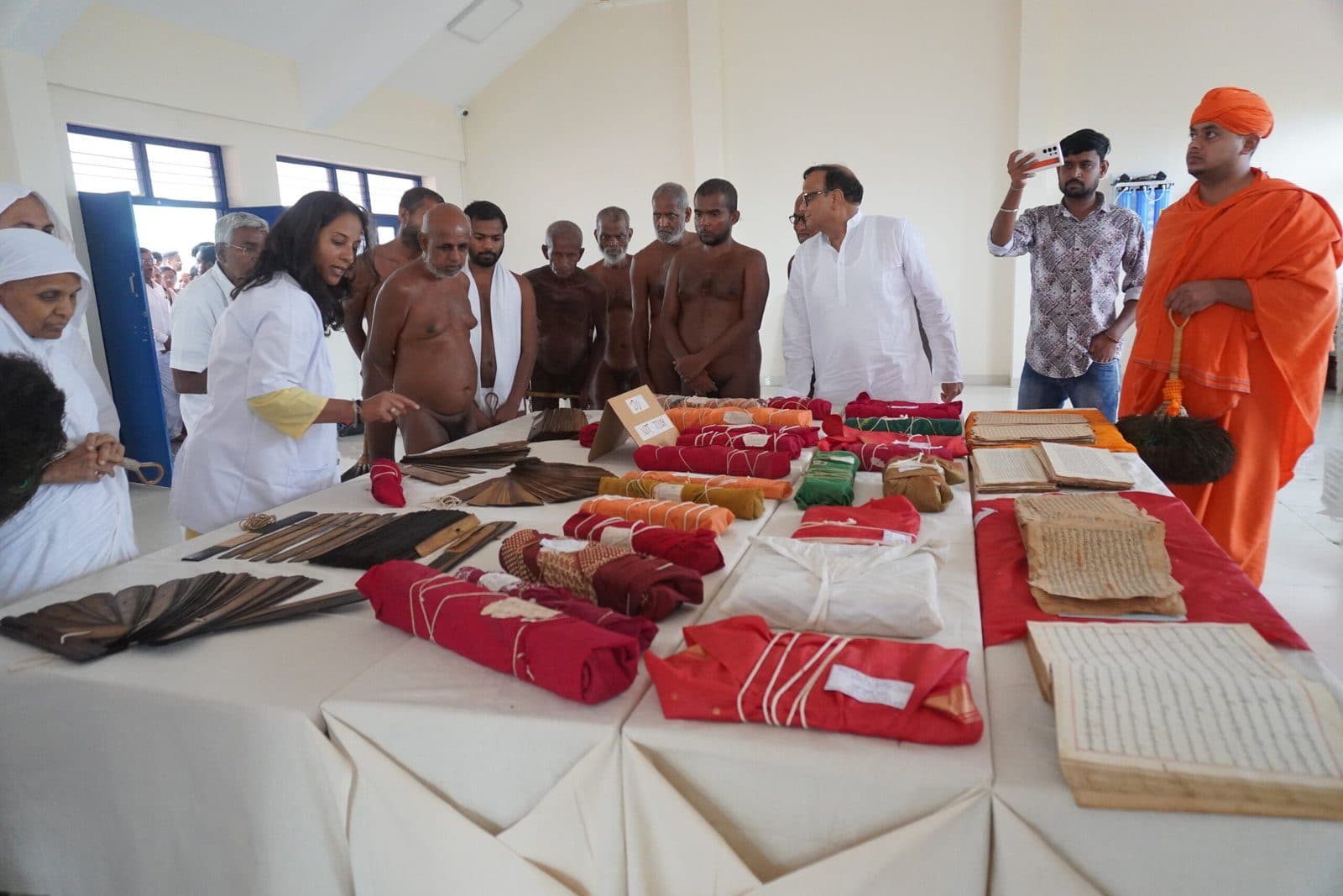 Munis and Foundation members around tables of palm-leaf bundles and cloth-wrapped manuscripts on red cloth.