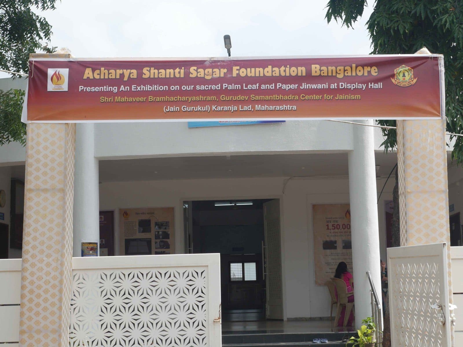 A building entrance under a maroon Foundation banner announcing an exhibition of palm-leaf and paper Jinvani.