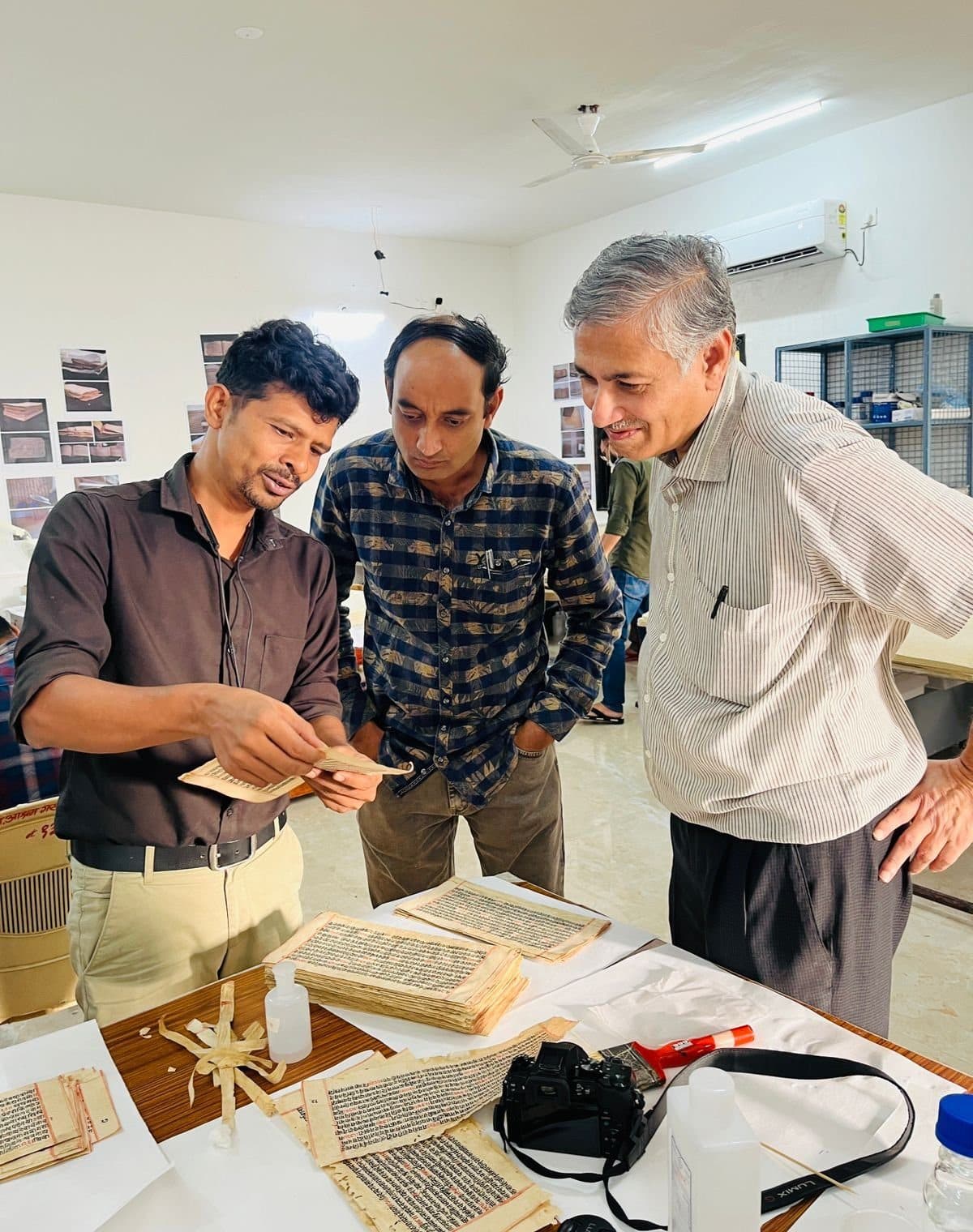 Three men examining handwritten folios spread on a table beside a camera.