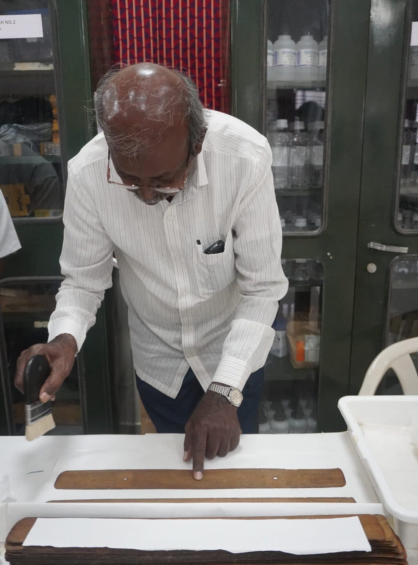 A man in a white shirt working on a palm leaf laid on a padded board.